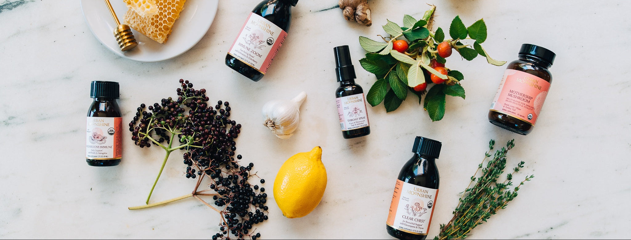 Four small amber glass bottles with black caps and pink labels are arranged on a white marble surface with natural ingredients including a lemon, garlic, elderberries, ginger, and herbs.